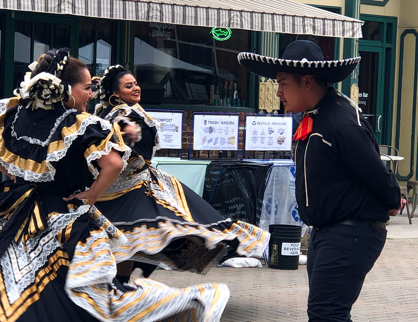 Dancers in traditional Mexican costumes perform on a street with recycling and compost bins in the background